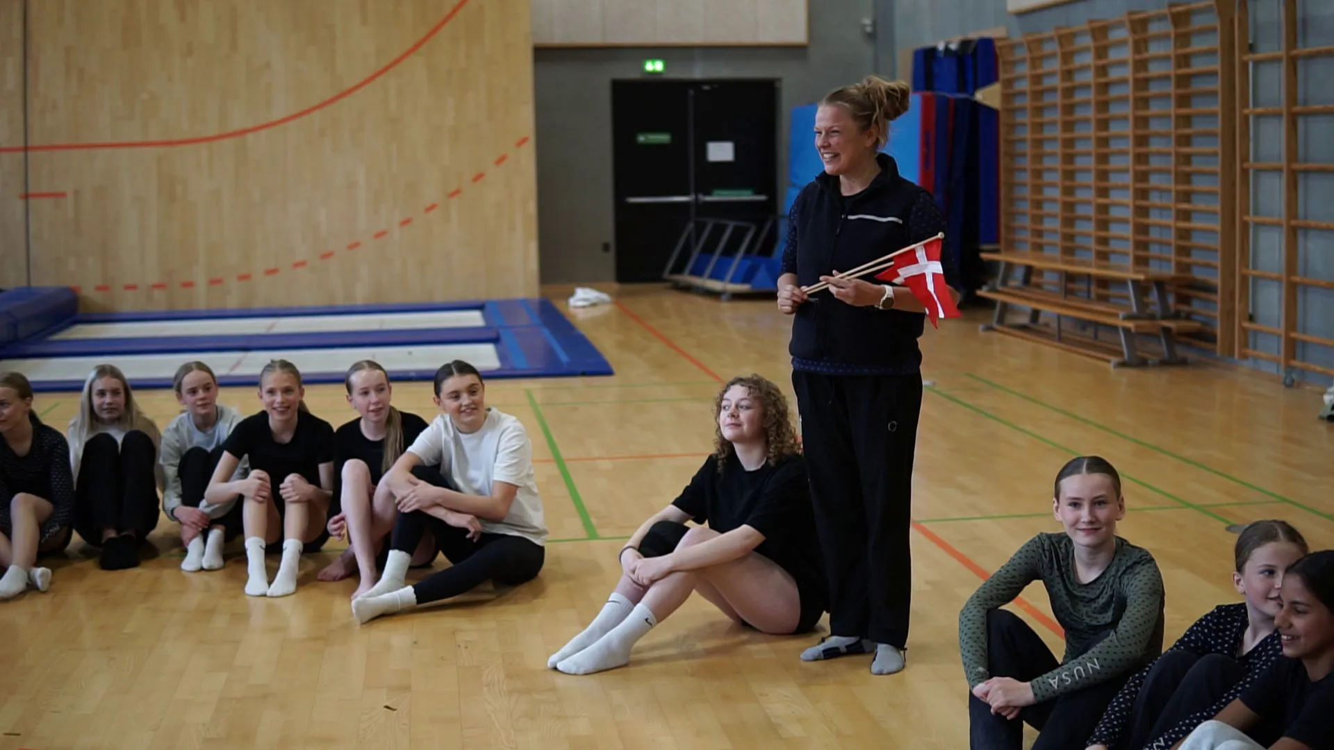 Instruktørkollega Sara Middelhede kommer ind i hallen med flag i hånden og overrasker Mathilde, mens gymnasterne sidder i rundkreds og ser måbende til.